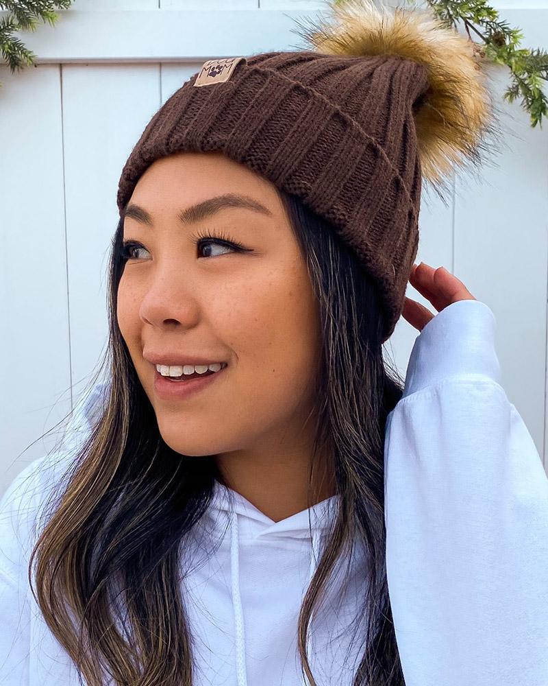 A young woman wearing a brown knit beanie with a puff ball on top that looks like a pomeranian's tail.